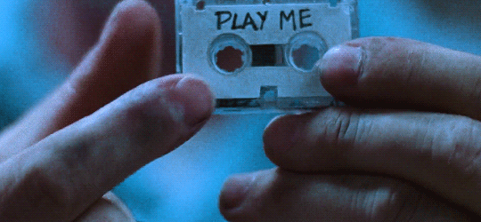 A pair of hands turning over a small video tape labeled "PLAY ME"